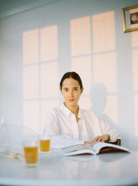 Woman poses indoors at a table with an open book and drinks, window light casting shadows on the white paneled wall behind her.