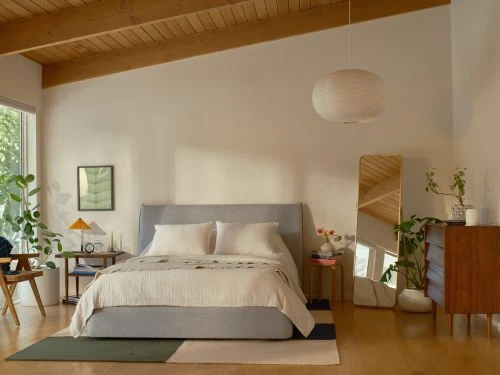 Modern bedroom interior with a grey bed, wooden dresser, potted plants, framed art, wooden floors, and natural light from the window.
