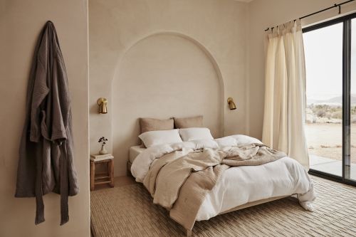 Minimalist bedroom interior with a white platform bed, soft beige walls, and a hanging bathrobe in a serene, naturally lit space.