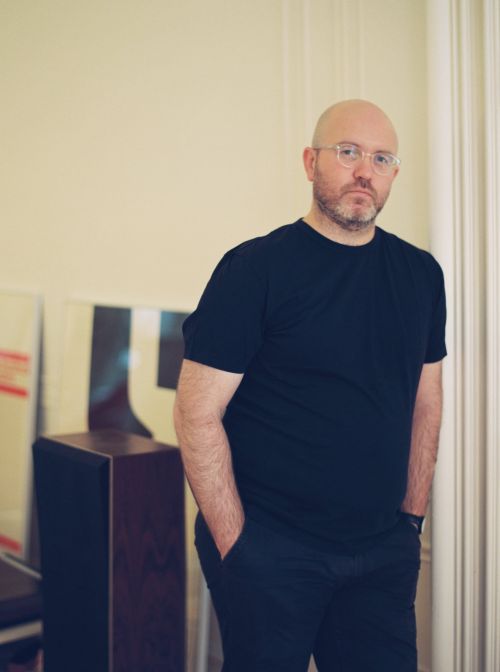 Indoor portrait of a bald adult male with a beard and glasses, wearing a dark T-shirt and pants, standing in front of artwork.