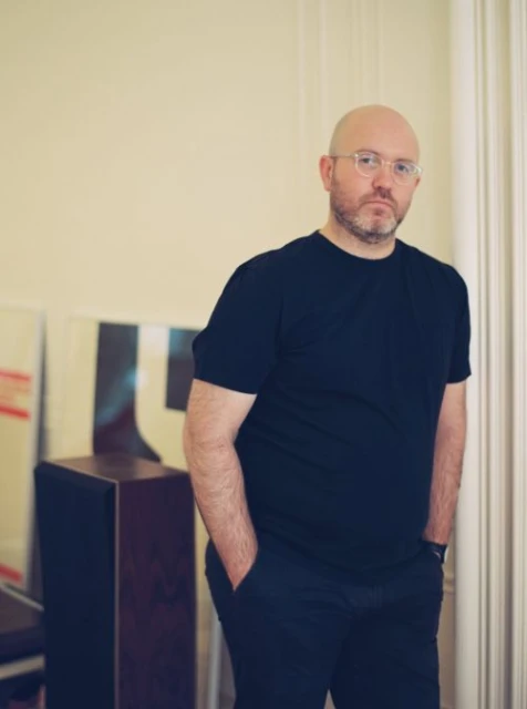 Indoor portrait of a bald adult male with a beard and glasses, wearing a dark T-shirt and pants, standing in front of artwork.