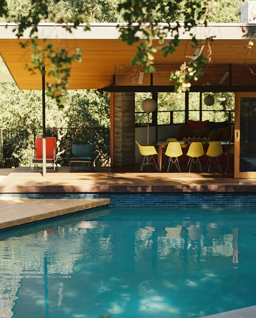 Modern house exterior with turquoise pool, open living space featuring yellow chairs, and natural light filtering through trees.