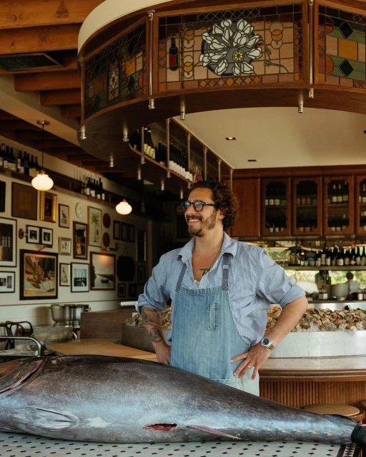 Adult male chef posing in a restaurant kitchen next to a large raw tuna, with wine bottles and framed art visible in the background.