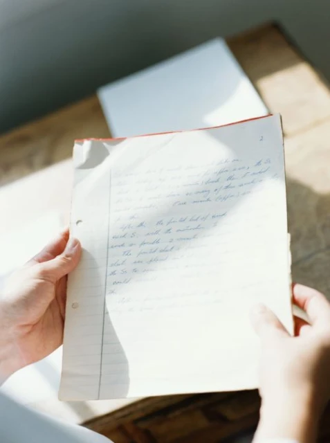 Person holds handwritten notes in a sunlit room, revealing details of the script, aged paper, and relaxed, academic atmosphere.