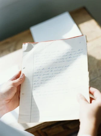 Person holds handwritten notes in a sunlit room, revealing details of the script, aged paper, and relaxed, academic atmosphere.