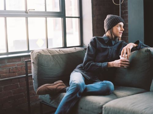 Woman sitting on a gray couch in a light-filled loft apartment wearing a beanie, jeans, and a dark shirt holding a cup.