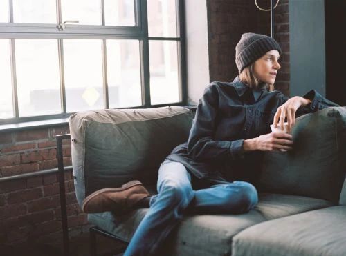 Woman sitting on a gray couch in a light-filled loft apartment wearing a beanie, jeans, and a dark shirt holding a cup.