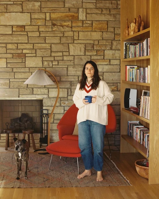 Woman holding a mug standing in a room with a dog, red chair, stone wall with fireplace, and wooden bookshelf. Warm, indoor lighting.