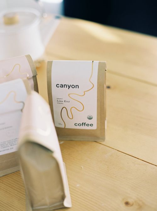 Close-up of Canyon Coffee bags on a light wood table with a white ceramic teapot blurred in the background. Food and beverage scene.
