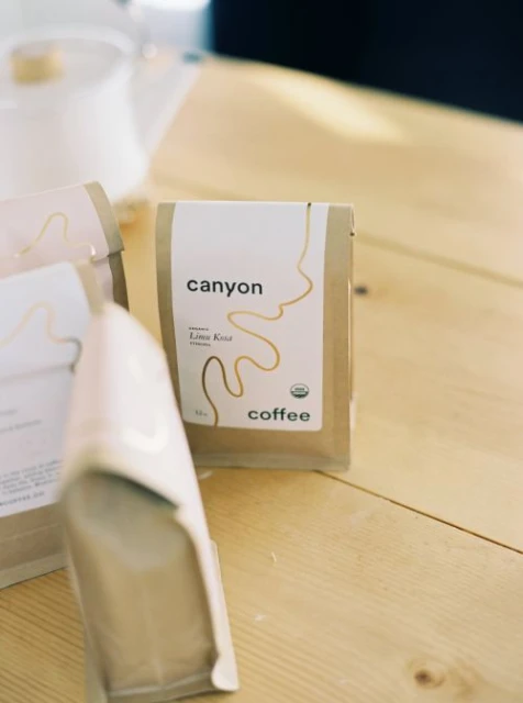Close-up of Canyon Coffee bags on a light wood table with a white ceramic teapot blurred in the background. Food and beverage scene.