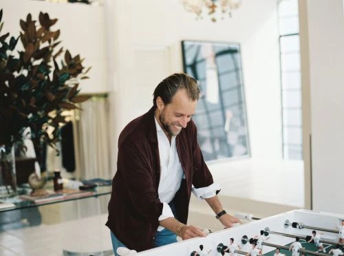 A man in a burgundy jacket smiles while playing foosball in a bright, modern living room with a large plant and artwork.