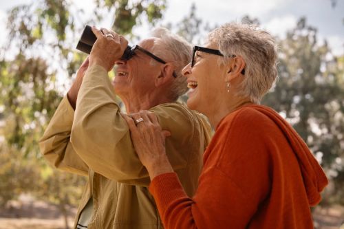 A happy elderly couple in an outdoor setting, with the man looking through binoculars and the woman smiling next to him.