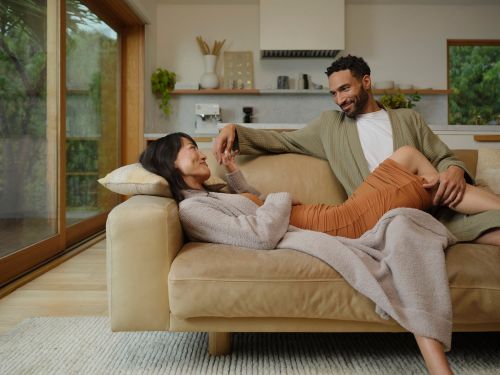 Couple relaxes on a beige couch in a bright living room. The woman is lying down while the man is sitting next to her, looking at her.