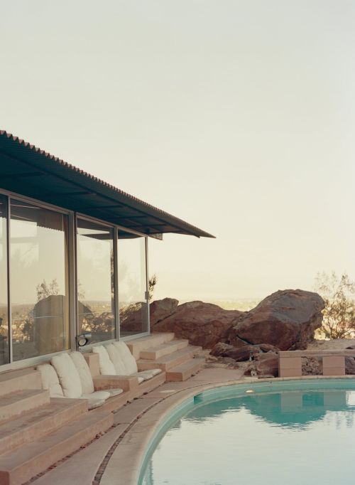 Exterior view of a mid-century modern house with a turquoise swimming pool, a concrete patio, and desert landscaping under a hazy sky.
