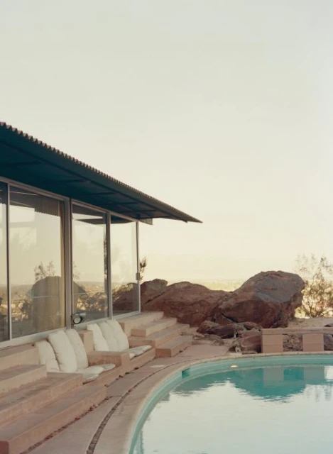Exterior view of a mid-century modern house with a turquoise swimming pool, a concrete patio, and desert landscaping under a hazy sky.