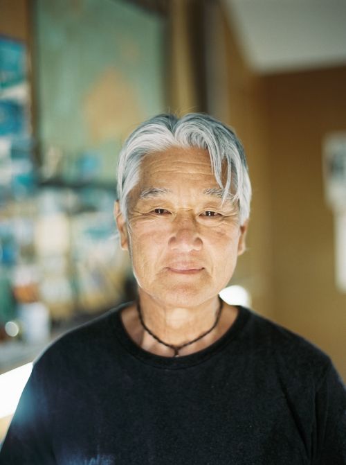Close-up portrait of an elderly Asian man with gray hair wearing a black shirt and necklace, posing indoors with a neutral facial expression.