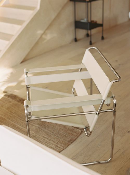 Minimalist modern chair with white canvas and tubular steel frame on a jute rug next to light wood steps in a sunlit room.