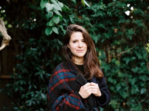 Portrait of a young adult woman in a red and blue plaid coat standing outside with dark green foliage in the background.