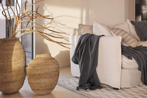 Interior decor featuring woven vases with branches next to a linen couch with a gray throw and striped rug in a neutral room.