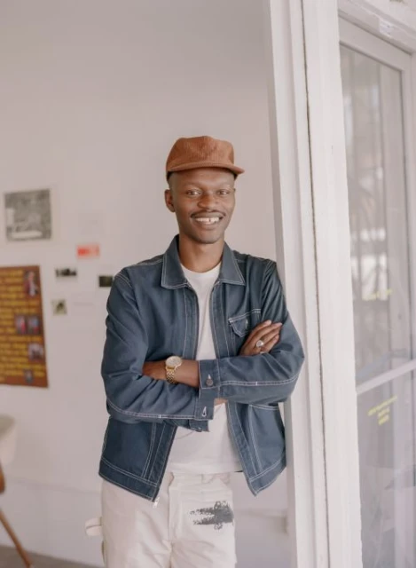 Portrait of a smiling Black man in a blue jacket and tan cap, arms crossed, leaning against a white doorframe in an indoor setting.