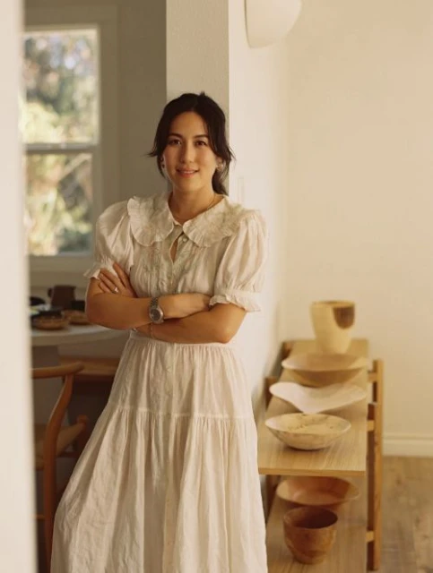 Portrait of a woman in a white dress with arms crossed, posing indoors near a wooden shelf filled with wooden bowls and vessels.