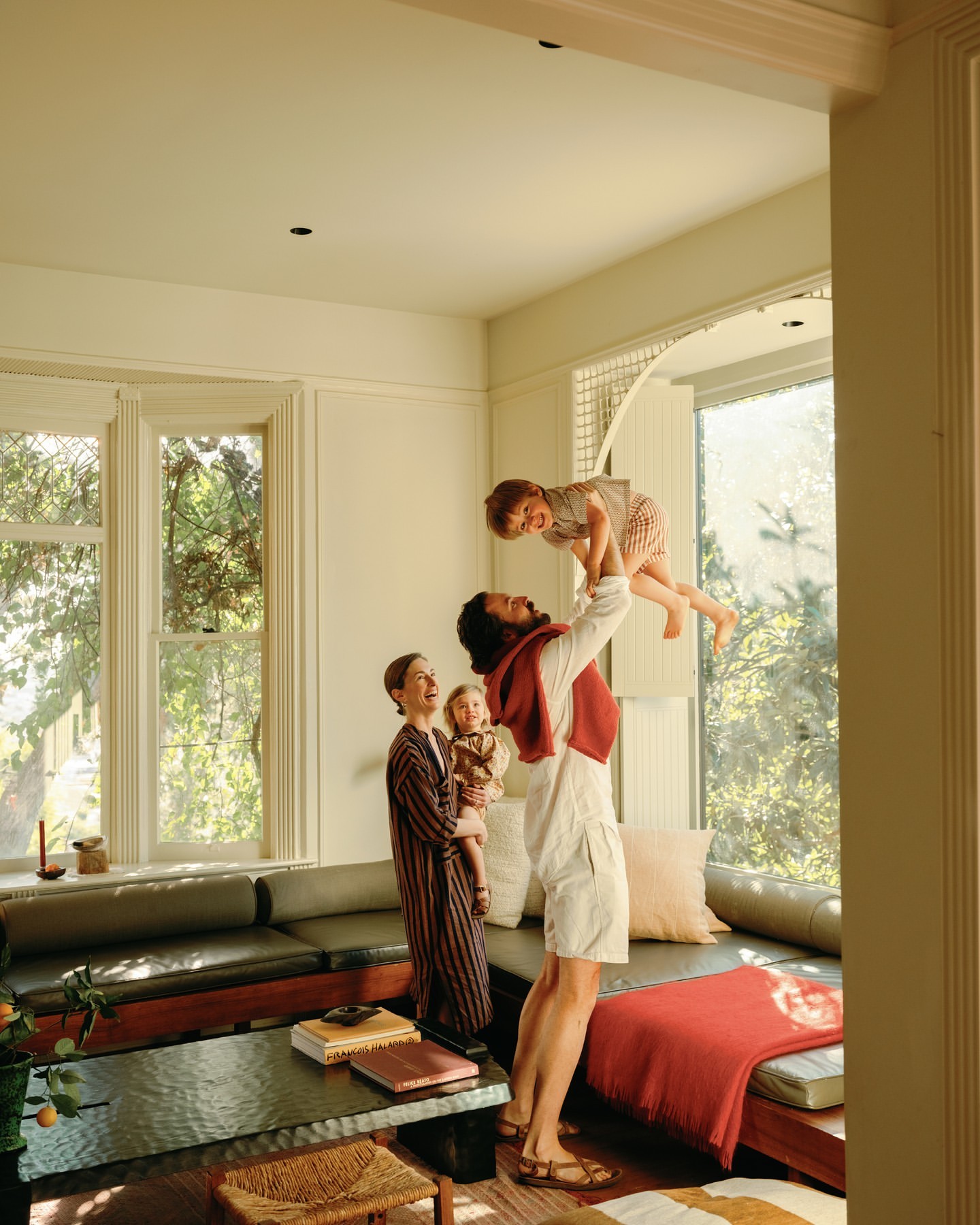 Happy family in brightly lit living room: father lifting young son in the air, mother holding toddler, modern interior design.