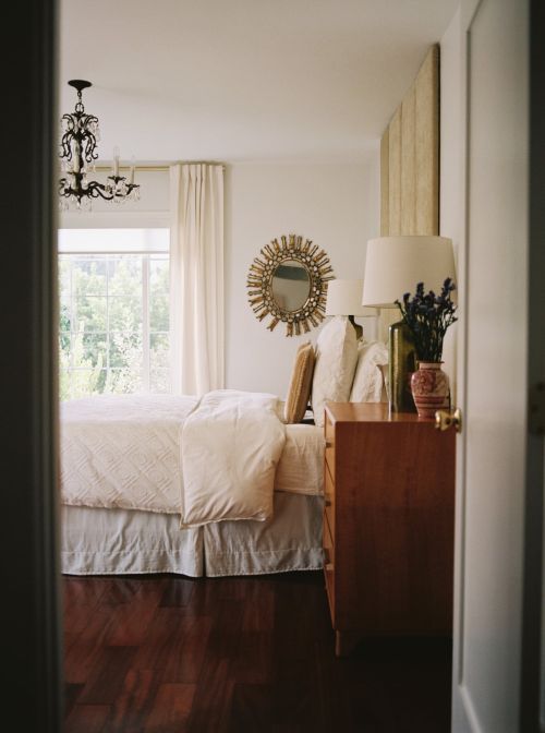Bedroom interior with a bed, wooden dresser, decorative mirror, chandelier, and window with natural light and dark wood floors.
