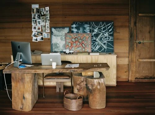 Photo of a modern wooden desk with an iMac, laptop, and woven basket in an indoor office space, illuminated by natural light.