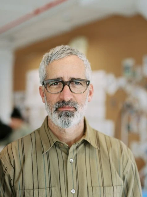 Portrait of an adult male with grey hair and beard wearing glasses and a button-down shirt in a bright, indoor environment.