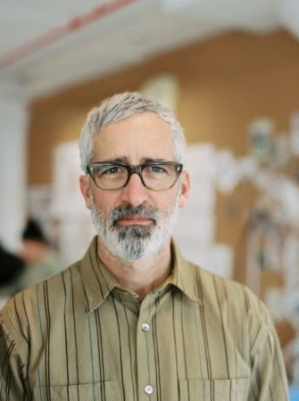 Portrait of an adult male with grey hair and beard wearing glasses and a button-down shirt in a bright, indoor environment.