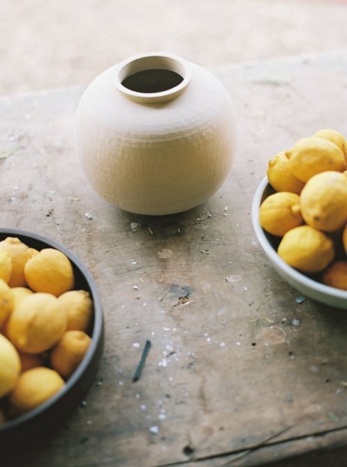 Still life photograph of a ceramic vase on a wooden surface with two bowls of bright yellow lemons arranged around it.
