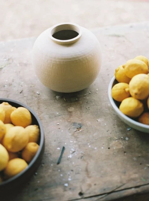 Still life photograph of a ceramic vase on a wooden surface with two bowls of bright yellow lemons arranged around it.
