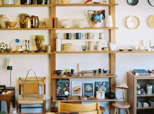 Wooden shelving unit displays pottery, mugs, books, and kitchenware in an indoor retail setting with natural light and a cozy, curated aesthetic.