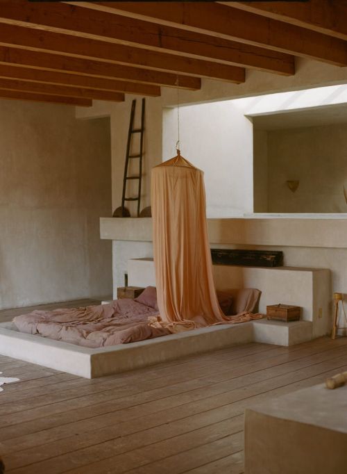 Minimalist bedroom with neutral tones featuring a raised platform bed, wooden floors, and draped fabric creating a serene atmosphere.