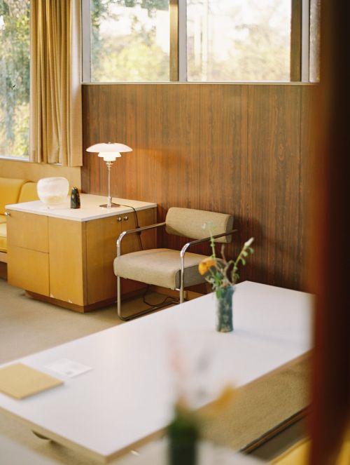 Mid-century modern interior with seating area: chair, table, and cabinet, plus a lamp, flowers, and view through windows.