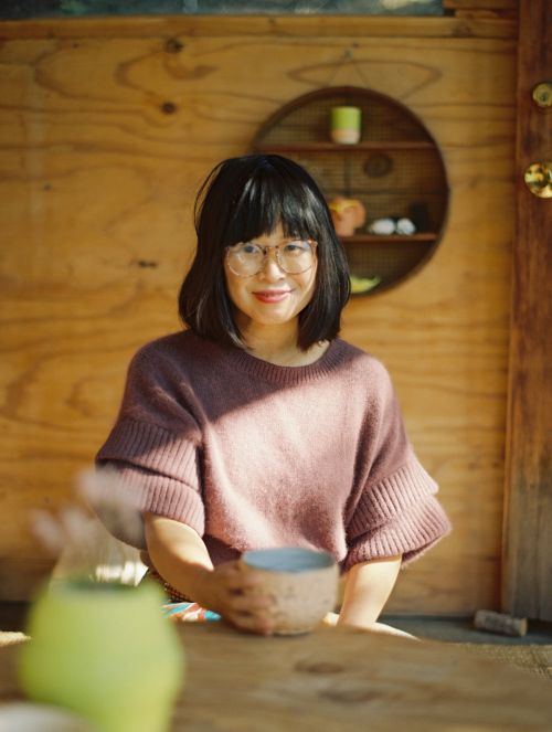 Indoor portrait of an adult woman with glasses, holding a ceramic cup, posing in front of a wooden wall and circular shelf.