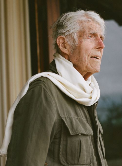 Elderly man in profile wearing a green jacket and white scarf stands indoors near a window with a neutral expression and natural light.