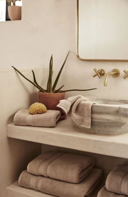 Bathroom interior features neatly stacked beige towels, a natural sponge, a potted plant, and a round sink with brass fixtures.
