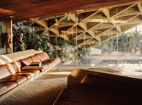 A mid-century modern living room features a brown leather sofa with geometric ceiling, glass wall, and lush greenery creating a tranquil atmosphere.