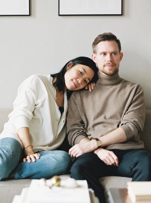 A young couple sits close together on a grey sofa with decorative books, light-colored walls, and framed prints in the background.