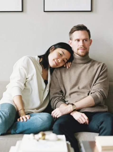 A young couple sits close together on a grey sofa with decorative books, light-colored walls, and framed prints in the background.