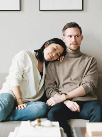 A young couple sits close together on a grey sofa with decorative books, light-colored walls, and framed prints in the background.