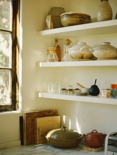 Warm-toned kitchen interior with natural light showcasing floating shelves with assorted pottery and glassware, along with stoneware cooking pots.