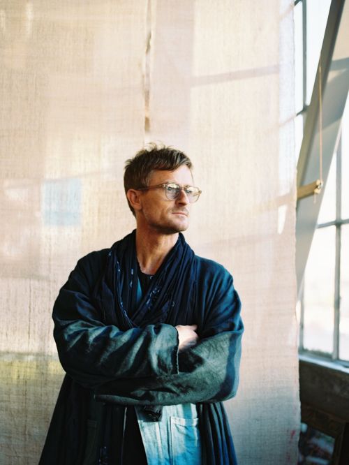 Portrait of an adult man wearing glasses and a navy robe, indoors with natural light, arms crossed, looking off to the side.