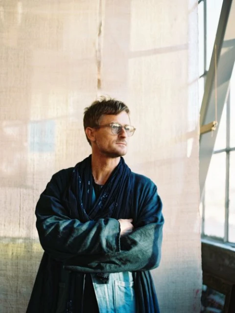 Portrait of an adult man wearing glasses and a navy robe, indoors with natural light, arms crossed, looking off to the side.