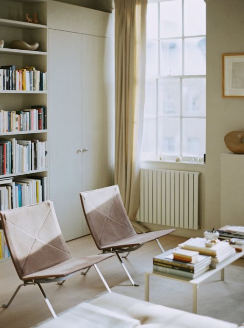 Interior shot of a living room featuring two sleek armchairs, a low coffee table stacked with books, and a tall bookshelf against the wall.