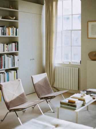 Interior shot of a living room featuring two sleek armchairs, a low coffee table stacked with books, and a tall bookshelf against the wall.