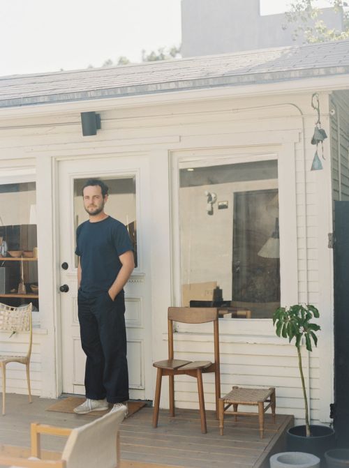 Adult male standing on a porch with chairs and potted plant, in front of a white house with windows and a door.