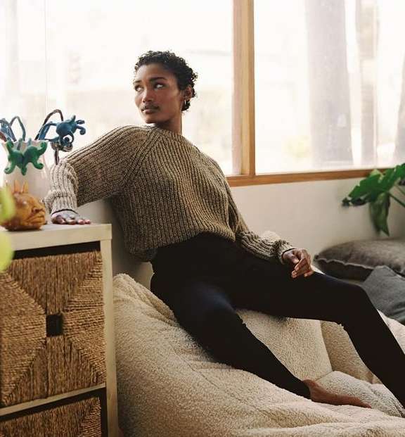 Adult female in a knitted sweater sits indoors on a beanbag chair near a window with natural light and indoor plants.