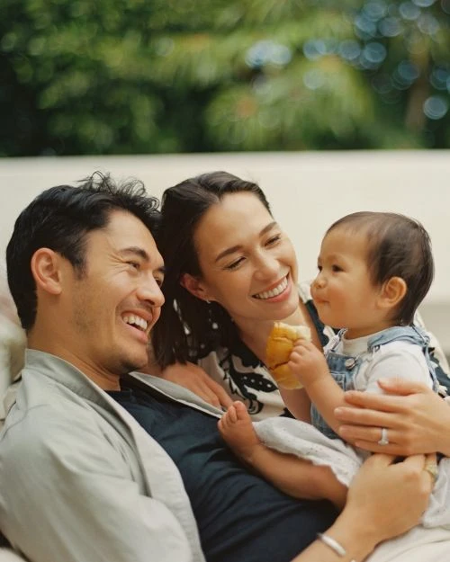 A family portrait captures a smiling man, woman, and baby together. The baby holds a bun as they are posed outdoors with blurred green foliage.